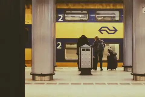 Pasajeros con maletas en un andén de la estación de tren de Schiphol junto a un tren de dos pisos amarillo y azul de NS