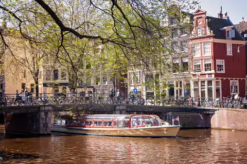 Barco turístico cubierto con techo de cristal pasando bajo un puente de piedra con bicicletas en un canal de Ámsterdam