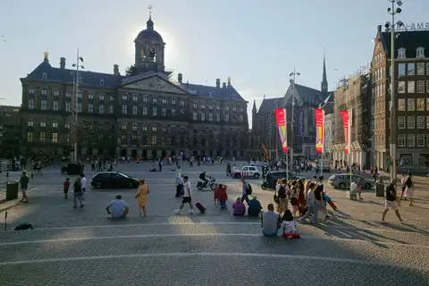Plaza Dam de Ámsterdam al atardecer con el Palacio Real, la torre de la Nieuwe Kerk al fondo y visitantes sentados y paseando