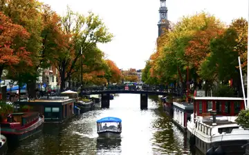 Canal de Ámsterdam en otoño con la torre de la Westerkerk, casas flotantes, un puente con bicicletas y un barco turístico cubierto