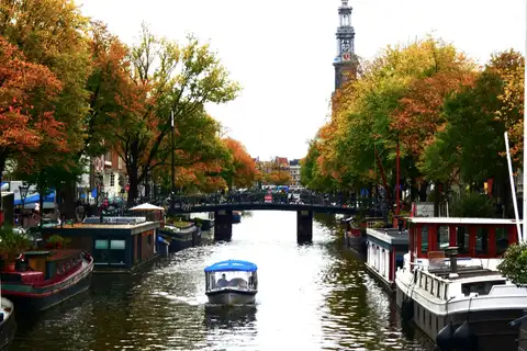 Canal de Ámsterdam en otoño con la torre de la Westerkerk, casas flotantes, un puente con bicicletas y un barco turístico cubierto
