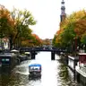 Canal de Ámsterdam en otoño con la torre de la Westerkerk, casas flotantes, un puente con bicicletas y un barco turístico cubierto