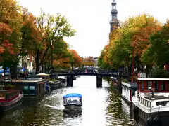 Canal de Ámsterdam en otoño con la torre de la Westerkerk, casas flotantes, un puente con bicicletas y un barco turístico cubierto