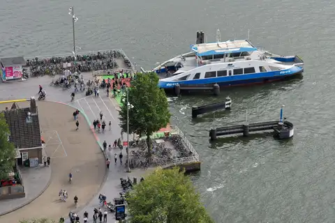 Vista aérea de un ferry azul y blanco de GVB atracado en un embarcadero del río IJ, con ciclistas y bicicletas aparcadas