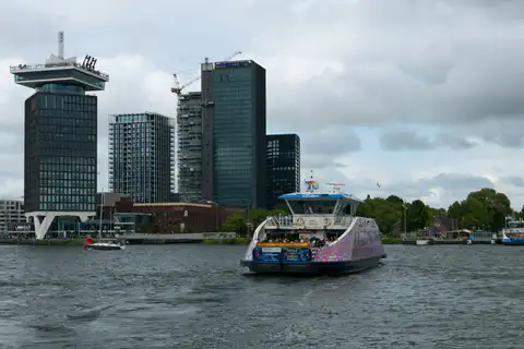 Ferry gratuito cruzando el río IJ con la torre A'DAM y edificios modernos de Ámsterdam Noord al fondo bajo un cielo nublado