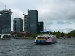 Ferry gratuito cruzando el río IJ con la torre A'DAM y edificios modernos de Ámsterdam Noord al fondo bajo un cielo nublado