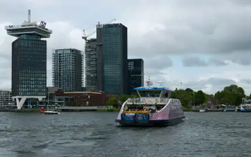Ferry gratuito cruzando el río IJ con la torre A'DAM y edificios modernos de Ámsterdam Noord al fondo bajo un cielo nublado