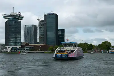Ferry gratuito cruzando el río IJ con la torre A'DAM y edificios modernos de Ámsterdam Noord al fondo bajo un cielo nublado