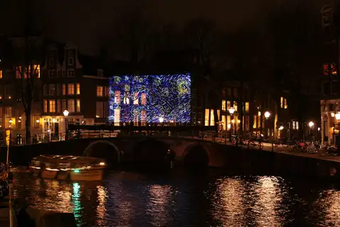Proyección luminosa inspirada en La noche estrellada sobre una fachada junto a un canal de Ámsterdam, con un puente de piedra y un barco en movimiento