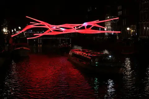 Barco de canal con techo de cristal pasando bajo una escultura luminosa roja sobre un canal de Ámsterdam de noche