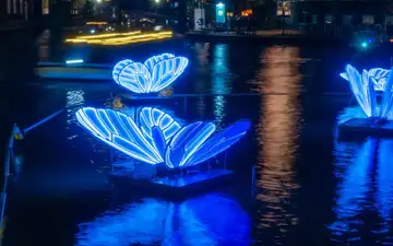 Mariposas luminosas azules flotando sobre un canal de Ámsterdam de noche, reflejadas en el agua, con edificios iluminados al fondo