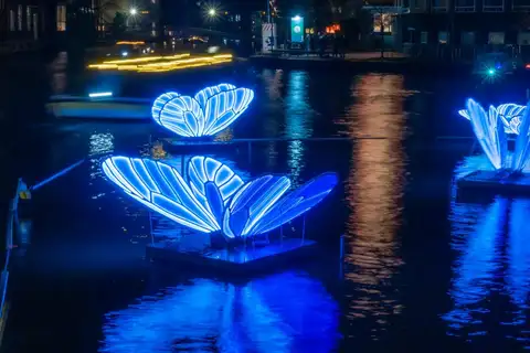 Mariposas luminosas azules flotando sobre un canal de Ámsterdam de noche, reflejadas en el agua, con edificios iluminados al fondo