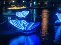 Mariposas luminosas azules flotando sobre un canal de Ámsterdam de noche, reflejadas en el agua, con edificios iluminados al fondo