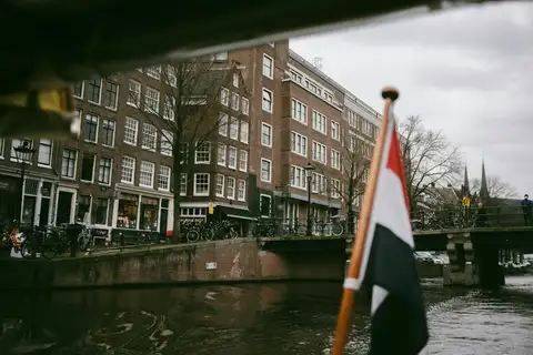 Bandera neerlandesa en un asta de madera junto a un canal de Ámsterdam, con casas de ladrillo, bicicletas y un puente al fondo