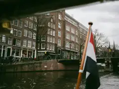 Bandera neerlandesa en un asta de madera junto a un canal de Ámsterdam, con casas de ladrillo, bicicletas y un puente al fondo