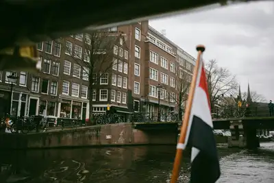 Bandera neerlandesa en un asta de madera junto a un canal de Ámsterdam, con casas de ladrillo, bicicletas y un puente al fondo