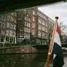 Bandera neerlandesa en un asta de madera junto a un canal de Ámsterdam, con casas de ladrillo, bicicletas y un puente al fondo