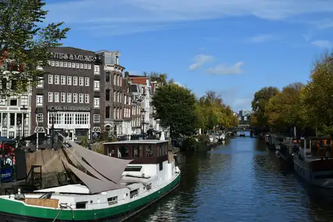 Canal del Jordaan en Ámsterdam con casas de ladrillo, casas flotantes amarradas y árboles de otoño bajo un cielo azul