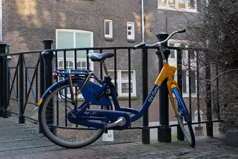 Bicicleta de alquiler azul y amarilla apoyada en la barandilla de hierro de un puente de Ámsterdam, con una fachada de ladrillo detrás
