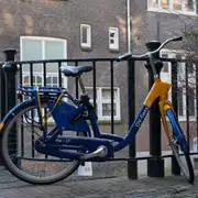 Bicicleta de alquiler azul y amarilla apoyada en la barandilla de hierro de un puente de Ámsterdam, con una fachada de ladrillo detrás