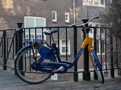 Bicicleta de alquiler azul y amarilla apoyada en la barandilla de hierro de un puente de Ámsterdam, con una fachada de ladrillo detrás