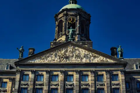 Torre del reloj con cúpula verde y frontón esculpido del Palacio Real de Ámsterdam, en la plaza Dam, bajo un cielo azul intenso