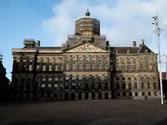 Fachada del Palacio Real de Ámsterdam con andamios en la cúpula, vista desde una plaza Dam vacía bajo un cielo azul con nubes