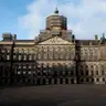 Fachada del Palacio Real de Ámsterdam con andamios en la cúpula, vista desde una plaza Dam vacía bajo un cielo azul con nubes