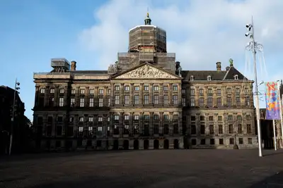 Fachada del Palacio Real de Ámsterdam con andamios en la cúpula, vista desde una plaza Dam vacía bajo un cielo azul con nubes
