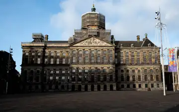Fachada del Palacio Real de Ámsterdam con andamios en la cúpula, vista desde una plaza Dam vacía bajo un cielo azul con nubes