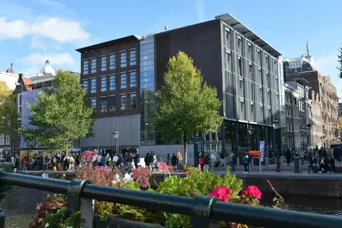 Edificio de ladrillo y cristal de la Casa de Ana Frank junto al canal Prinsengracht, con visitantes haciendo cola y flores en primer plano