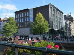 Edificio de ladrillo y cristal de la Casa de Ana Frank junto al canal Prinsengracht, con visitantes haciendo cola y flores en primer plano