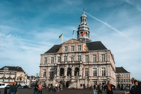 Ayuntamiento barroco de piedra con torre con reloj en la plaza Markt de Maastricht, con peatones y ciclistas bajo un cielo azul con nubes