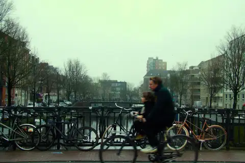 Padre y niño en bicicleta con abrigo cruzando un puente sobre un canal, con árboles sin hojas y niebla gris de invierno en Ámsterdam