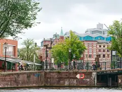 Ciclistas cruzando un puente de ladrillo sobre un canal de Ámsterdam bajo un cielo nublado, con fachadas de ladrillo y árboles verdes al fondo