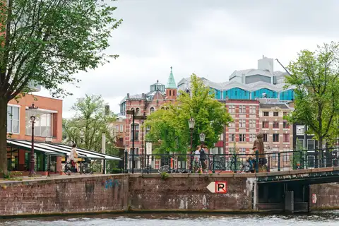 Ciclistas cruzando un puente de ladrillo sobre un canal de Ámsterdam bajo un cielo nublado, con fachadas de ladrillo y árboles verdes al fondo