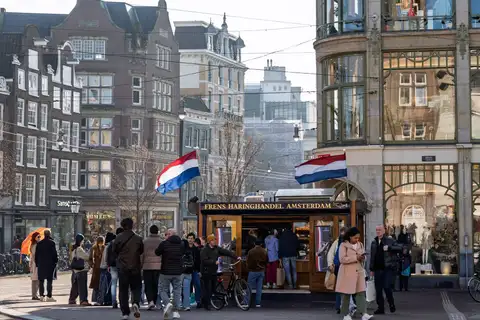 Gente haciendo cola en un puesto de arenque de madera con dos banderas neerlandesas ondeando, entre edificios de ladrillo de Ámsterdam