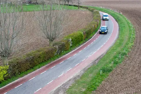 Vista aérea de una carretera rural curva con carriles bici rojos a ambos lados y dos coches con matrícula neerlandesa entre campos llanos