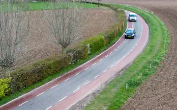 Vista aérea de una carretera rural curva con carriles bici rojos a ambos lados y dos coches con matrícula neerlandesa entre campos llanos