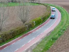 Vista aérea de una carretera rural curva con carriles bici rojos a ambos lados y dos coches con matrícula neerlandesa entre campos llanos
