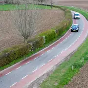 Vista aérea de una carretera rural curva con carriles bici rojos a ambos lados y dos coches con matrícula neerlandesa entre campos llanos