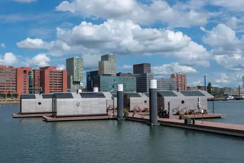 Casas flotantes modulares de madera amarradas a un muelle en un puerto de Rotterdam, con bloques de ladrillo y torres de viviendas y oficinas al fondo