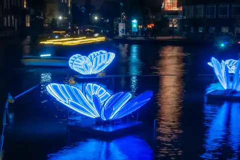 Mariposas gigantes de luz azul flotando sobre un canal de Ámsterdam de noche, con reflejos en el agua, en el Amsterdam Light Festival