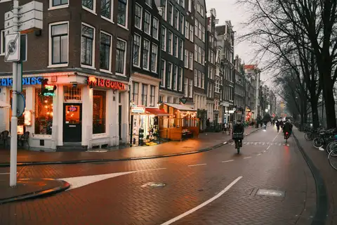Calle de ladrillo de Ámsterdam al anochecer, con ciclistas bajo los árboles y fachadas estrechas de ladrillo con escaparates iluminados