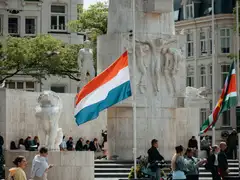 Bandera neerlandesa ondeando junto al Monumento Nacional de la plaza Dam, con relieves de piedra, un león y gente sentada en las escaleras