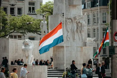 Bandera neerlandesa ondeando junto al Monumento Nacional de la plaza Dam, con relieves de piedra, un león y gente sentada en las escaleras