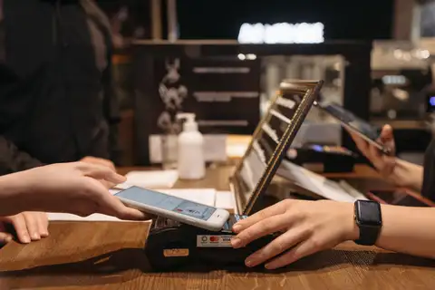Manos acercando un teléfono móvil al datáfono contactless sobre el mostrador de madera de un café