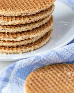 Pila de stroopwafels dorados sobre un plato blanco, con migas y otro stroopwafel apoyado sobre un paño azul