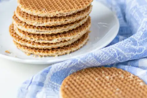 Pila de stroopwafels dorados sobre un plato blanco, con migas y otro stroopwafel apoyado sobre un paño azul