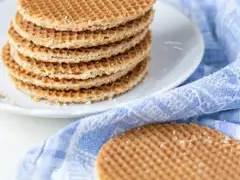 Pila de stroopwafels dorados sobre un plato blanco, con migas y otro stroopwafel apoyado sobre un paño azul